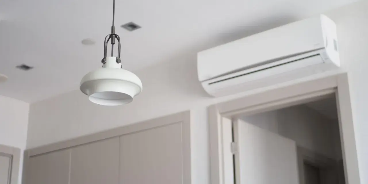 Biała klimatyzacja ścienna nad drzwiami, obok lampy wiszącej.