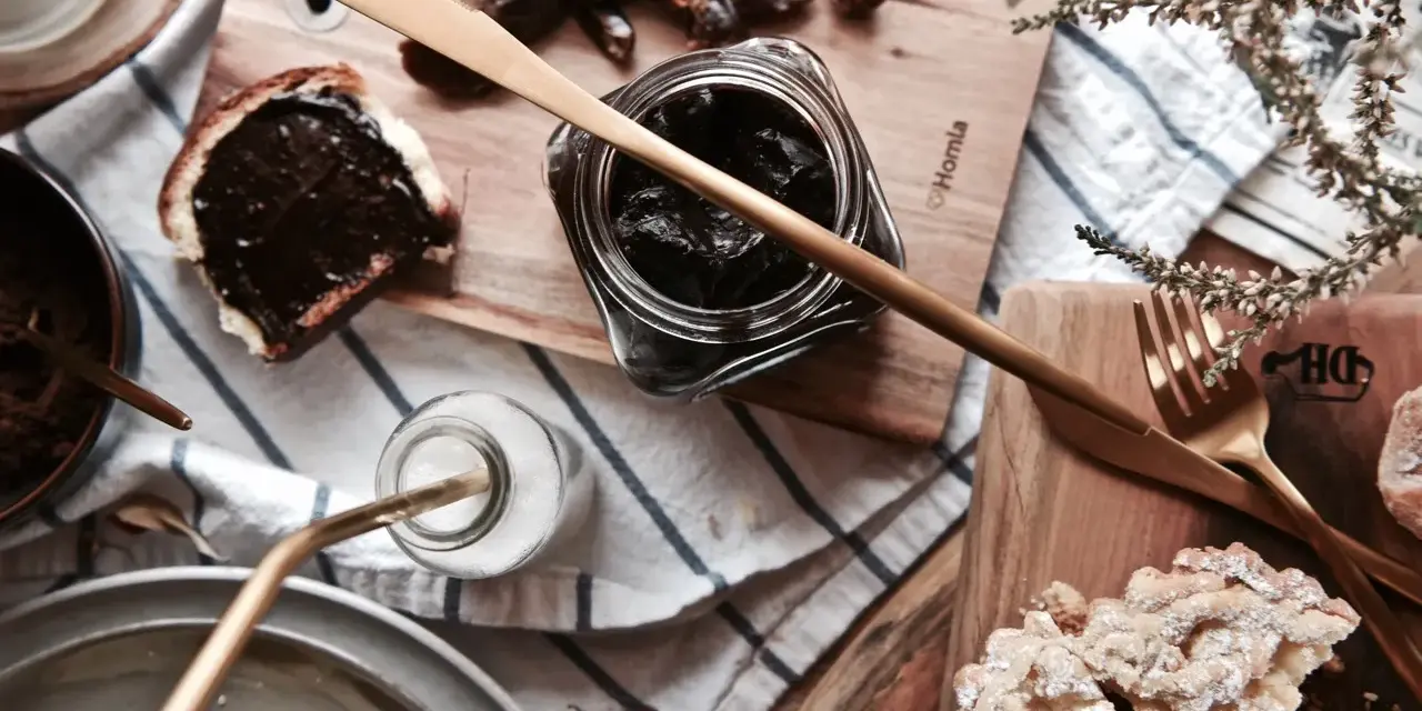 Słodkie śniadanie: kromka chleba z kremem czekoladowym, słoiczek z ciemnym dżemem, daktyle i ciastka. Idealna nutella bez orzechów!