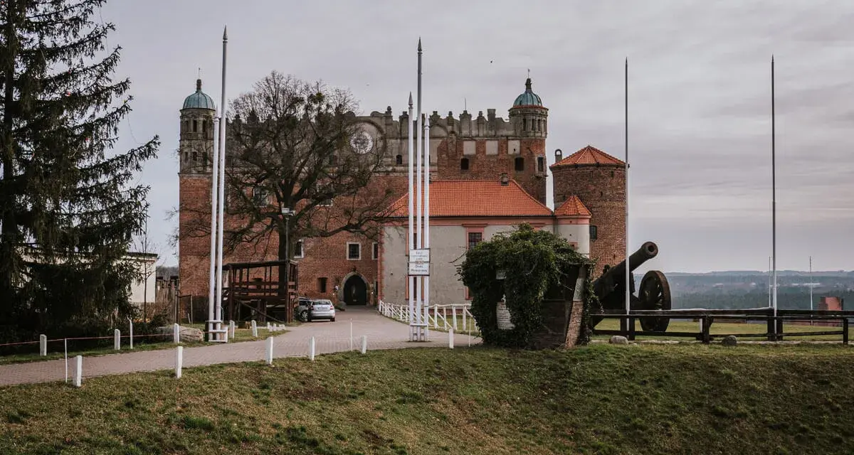 Zamek Golub-Dobrzyń zaprasza na zwiedzanie. Potężna twierdza z armatą na dziedzińcu, otoczona zielenią i drzewami.
