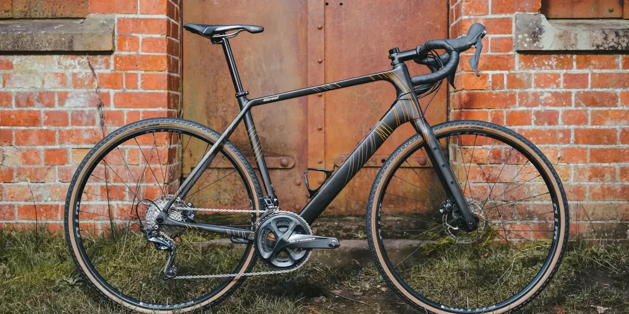A sleek black Salsa Warroad gravel bike with orange accents stands against a rustic brick wall and weathered metal door.
