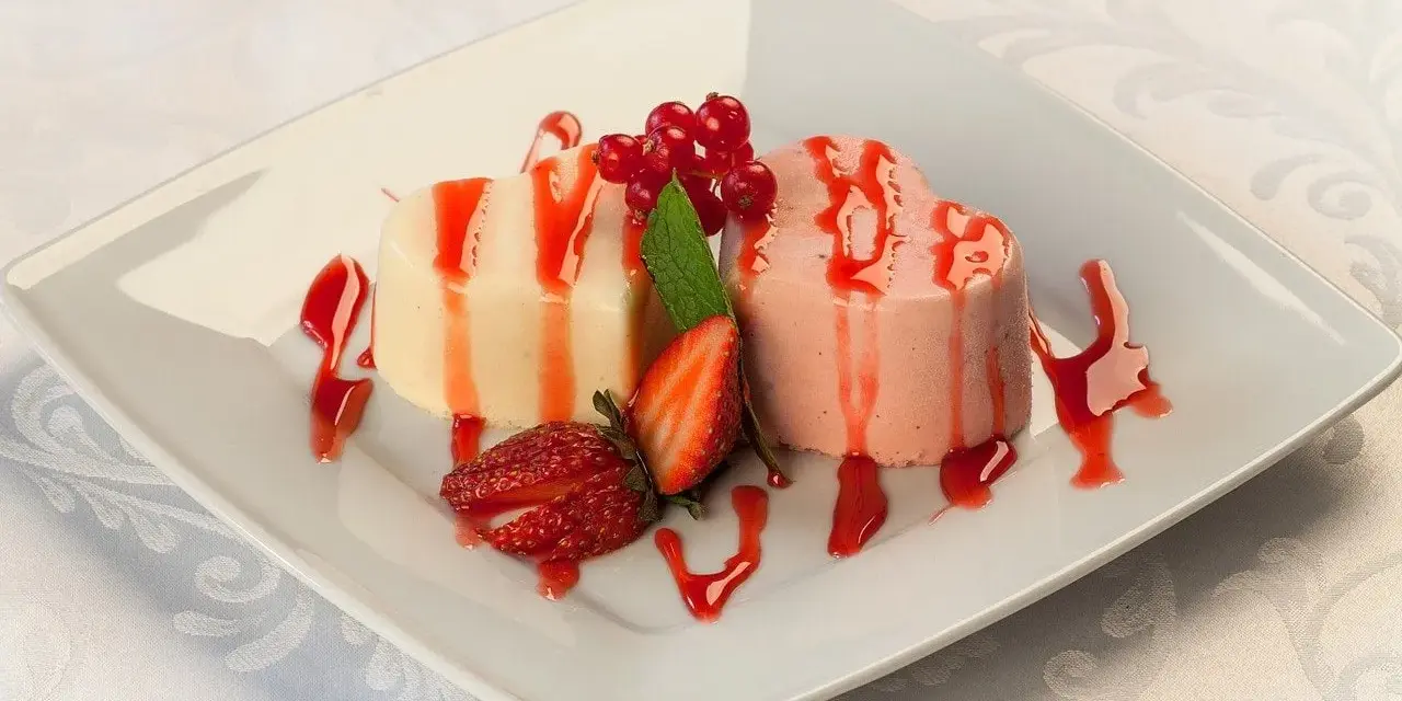 Dwie panna cotta w kształcie serc, polane sosem truskawkowym, ozdobione owocami i miętą.