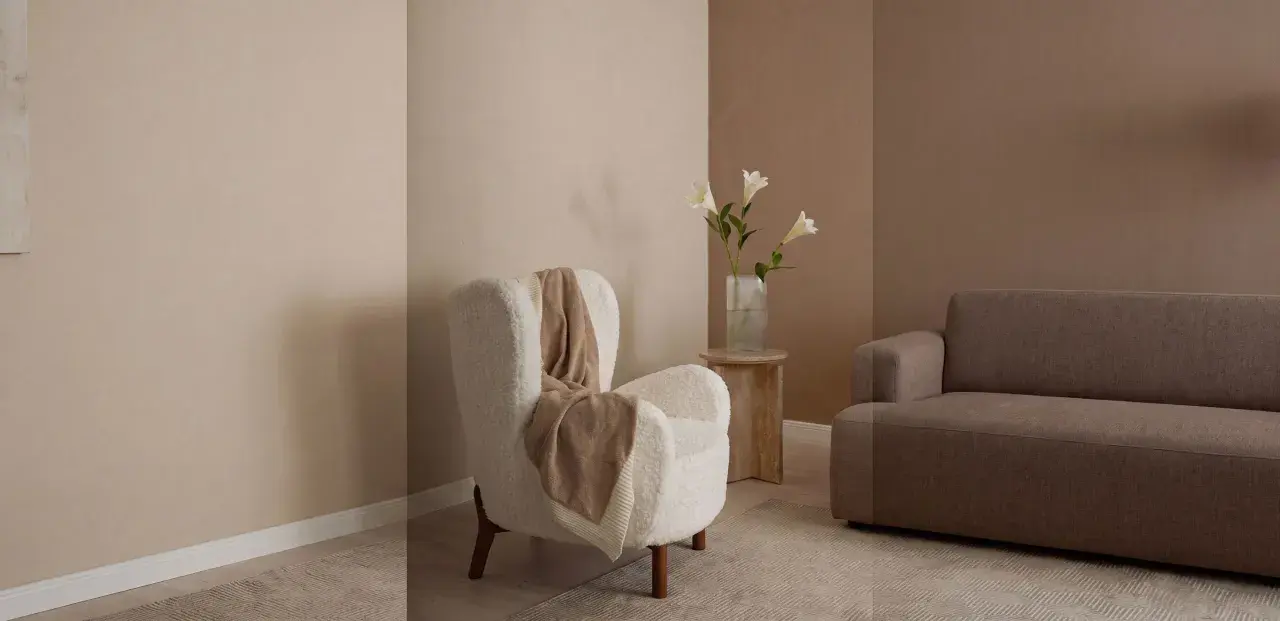 Gemütliches Wohnzimmer mit beigen und braunen Wandfarben. Ein flauschiger Sessel mit Decke und ein Sofa laden zum Entspannen ein.