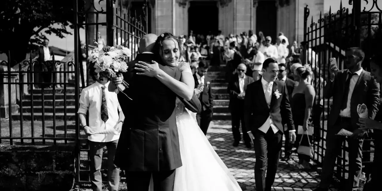Un couple s'embrasse devant une église, entouré d'invités. Un jeune garçon tient un bouquet. Ce moment est le fruit d'un savant **rétroplanning mariage**.
