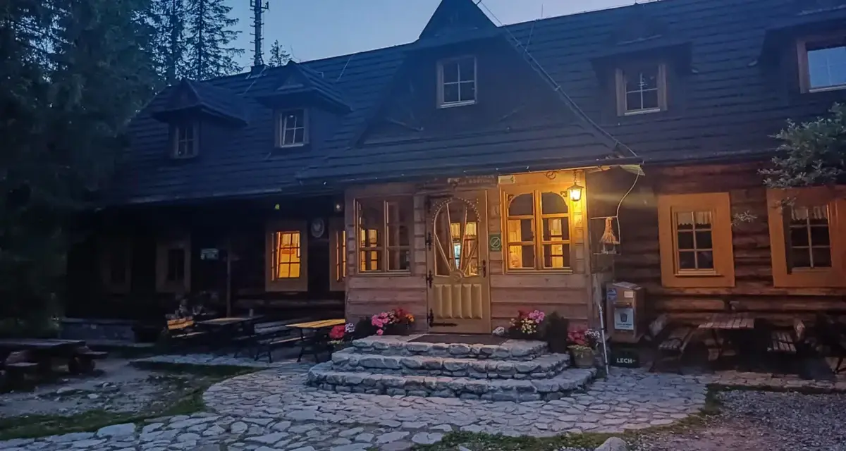 Drewniana chata w górach, oświetlona ciepłym światłem, to idealna baza pod roztokami. Kamienna ścieżka prowadzi do wejścia.