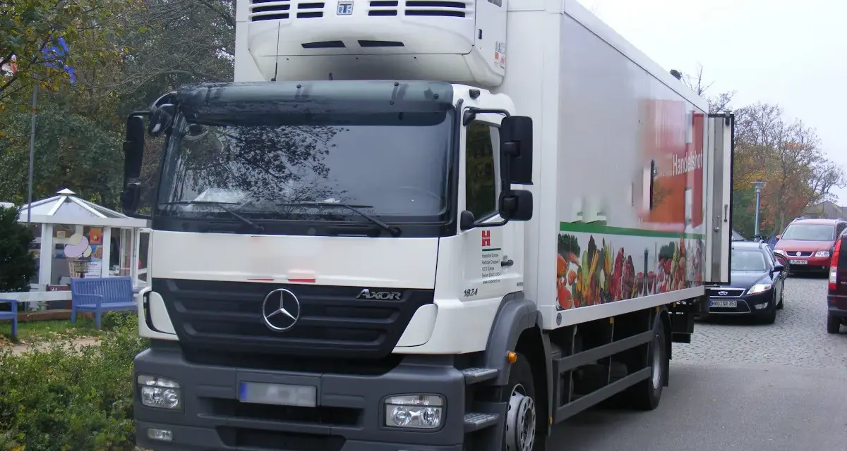 Biały Mercedes Axor z chłodnią, ozdobiony grafiką warzyw, gotowy do dostawy świeżych produktów.