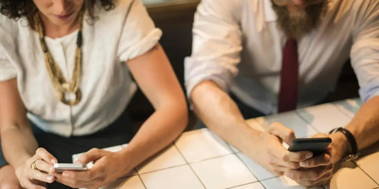 Zwei Personen sitzen an einem Tisch und schauen auf ihre Smartphones.
