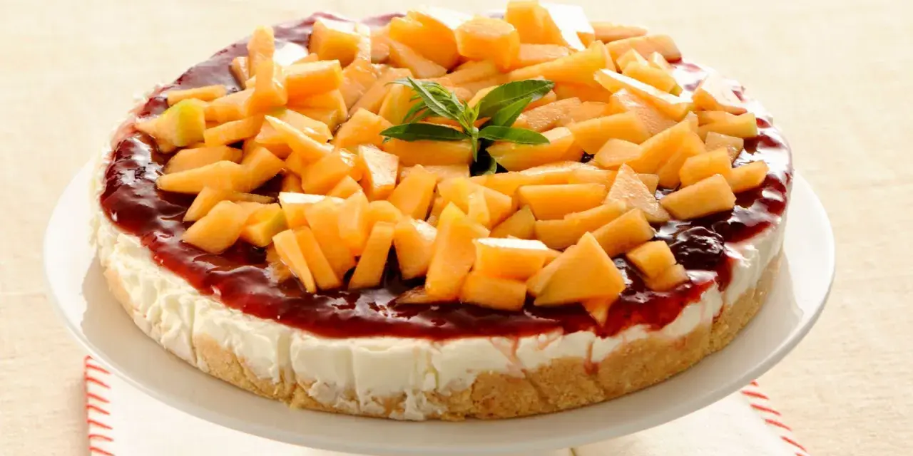 Deliziosa cheesecake al melone, con topping di frutta fresca e salsa rossa, su un piatto bianco.