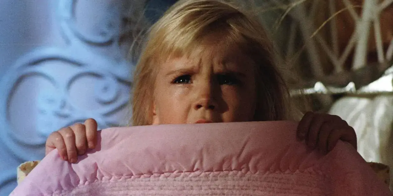 Bambina spaventata si nasconde sotto una coperta rosa, gli occhi sgranati in un'espressione di puro horror psicologico.