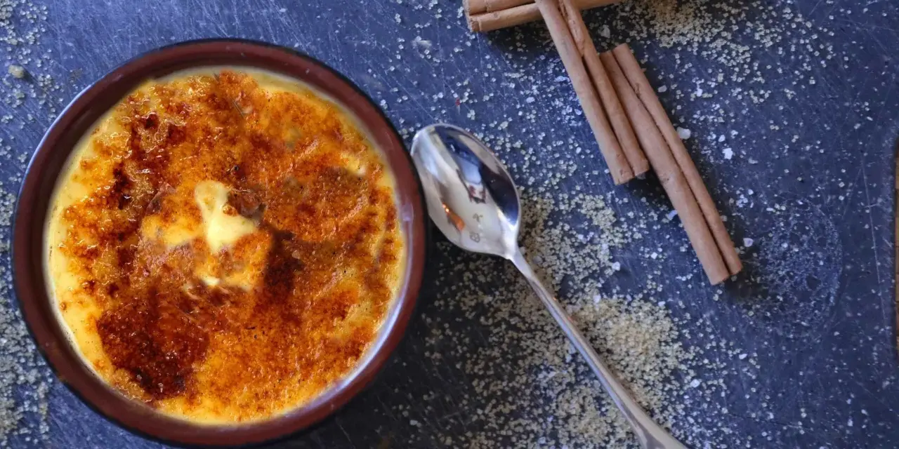 Cremige Crema Catalana mit karamellisierter Zuckerkruste, daneben Zimtstangen und Zucker.