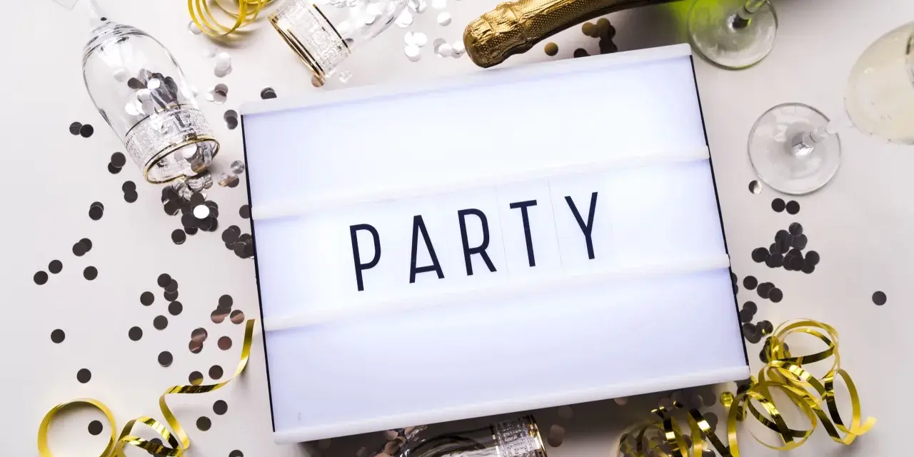 Świętowanie zaczyna się od zaproszeń! Na stole leżą kieliszki, szampan i konfetti, a tablica świetlna głosi "PARTY".