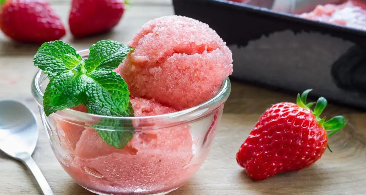 Erfrischendes Erdbeer-Sorbet-Eis in einer Glasschale, garniert mit Minze. Daneben liegen frische Erdbeeren.