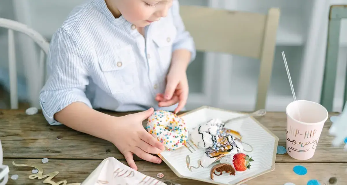 En pojke med en randig skjorta tar en donut med strössel. Det är en festlig dukning med konfetti och en mugg med texten "Hip Hip Hooray". En härlig stund av burken lek.