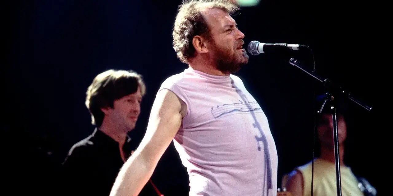 Joe Cocker śpiewa na scenie, a jego charakterystyczne utwory niosą się po sali. W tle widać innych muzyków.