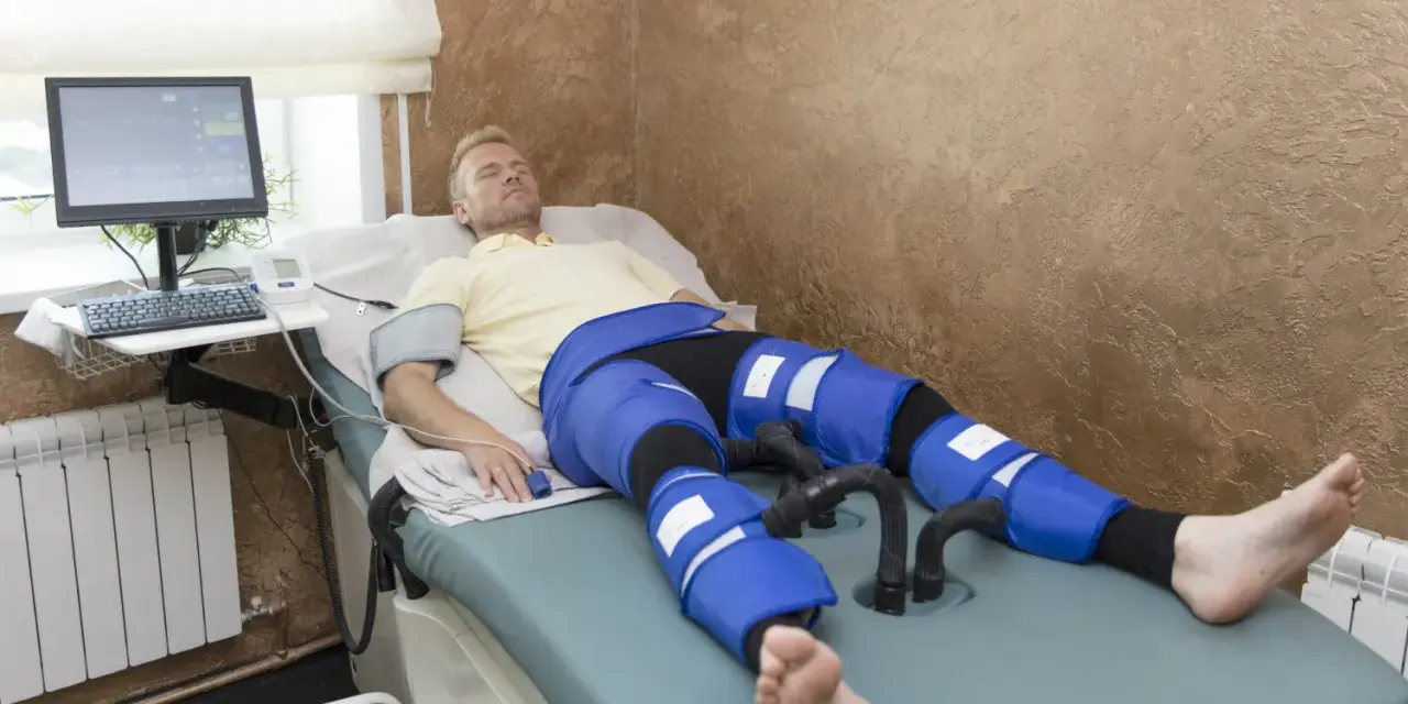 Mężczyzna leży na łóżku terapeutycznym, podłączony do aparatury do limfodrenażu. Widoczne są jego nogi w specjalnych mankietach, które mają zapewnić widoczne limfodrenaż efekty.
