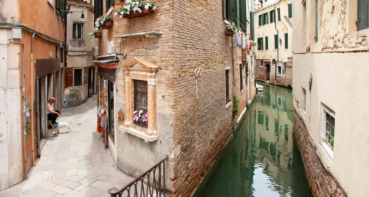 Un vicolo stretto e un canale a Venezia, con una donna e un cane seduti, tra i tanti **Venezia posti da visitare**.