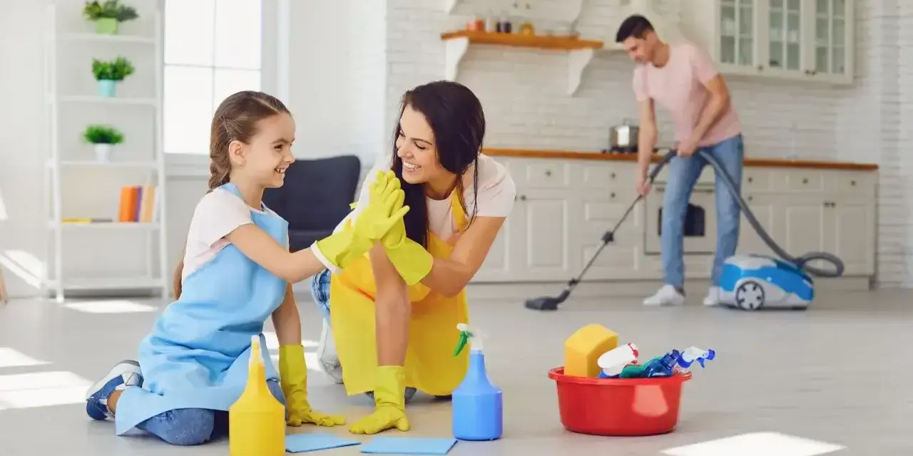 Mama i córka świętują wykonane obowiązki 9 latka w domu. Tata odkurza w tle.