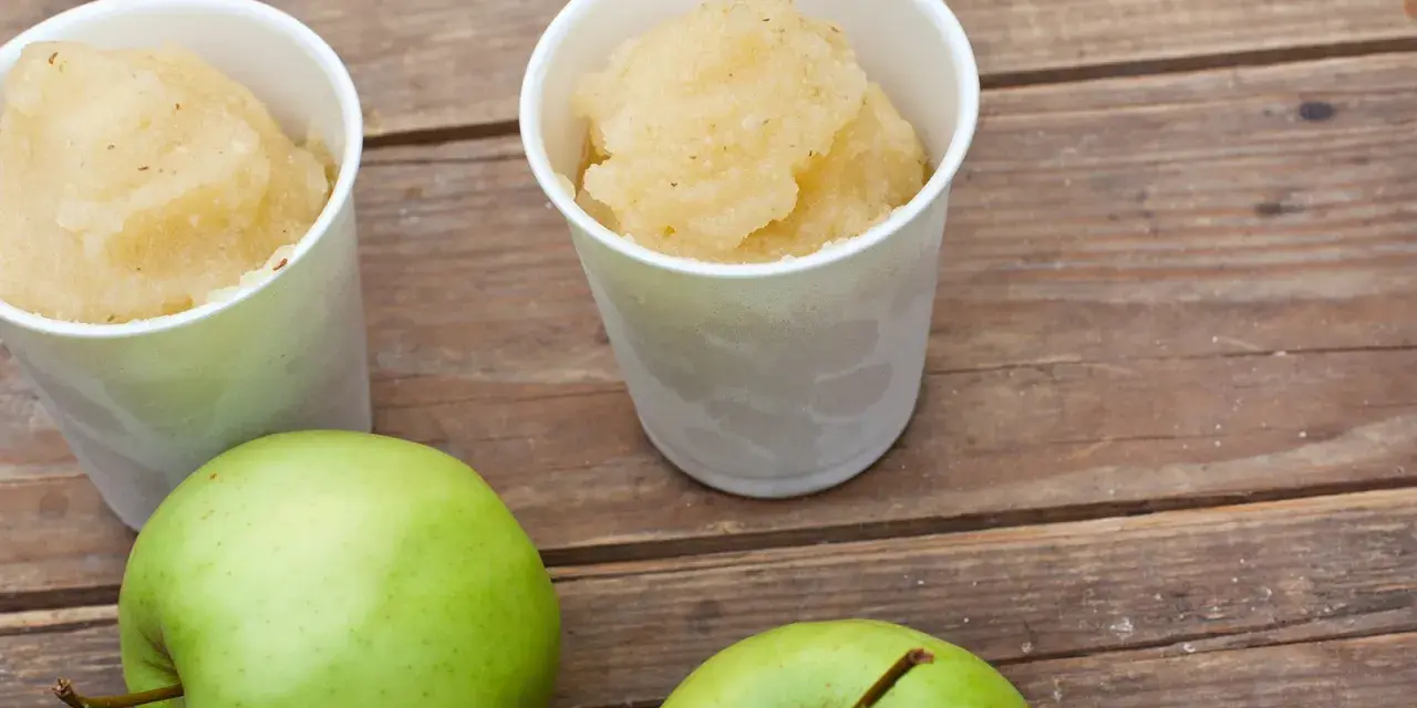 Zwei Becher mit erfrischendem Apfel Sorbet stehen neben zwei grünen Äpfeln auf einer Holzoberfläche.