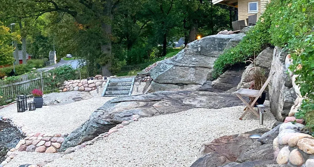 En stenlagd uteplats med grusgångar, stenmurar och trappor som leder upp mot ett hus. En inspiration för en naturlig och robust utomhusmiljö.