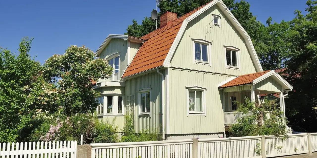 Ljusgrönt trähus med tegeltak och vit staket. En vacker trädgård med blommande buskar. Planerar att byta fasad för att fräscha upp huset.