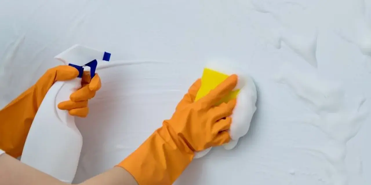 Manos con guantes naranjas rociando y limpiando paredes con espuma y esponja.