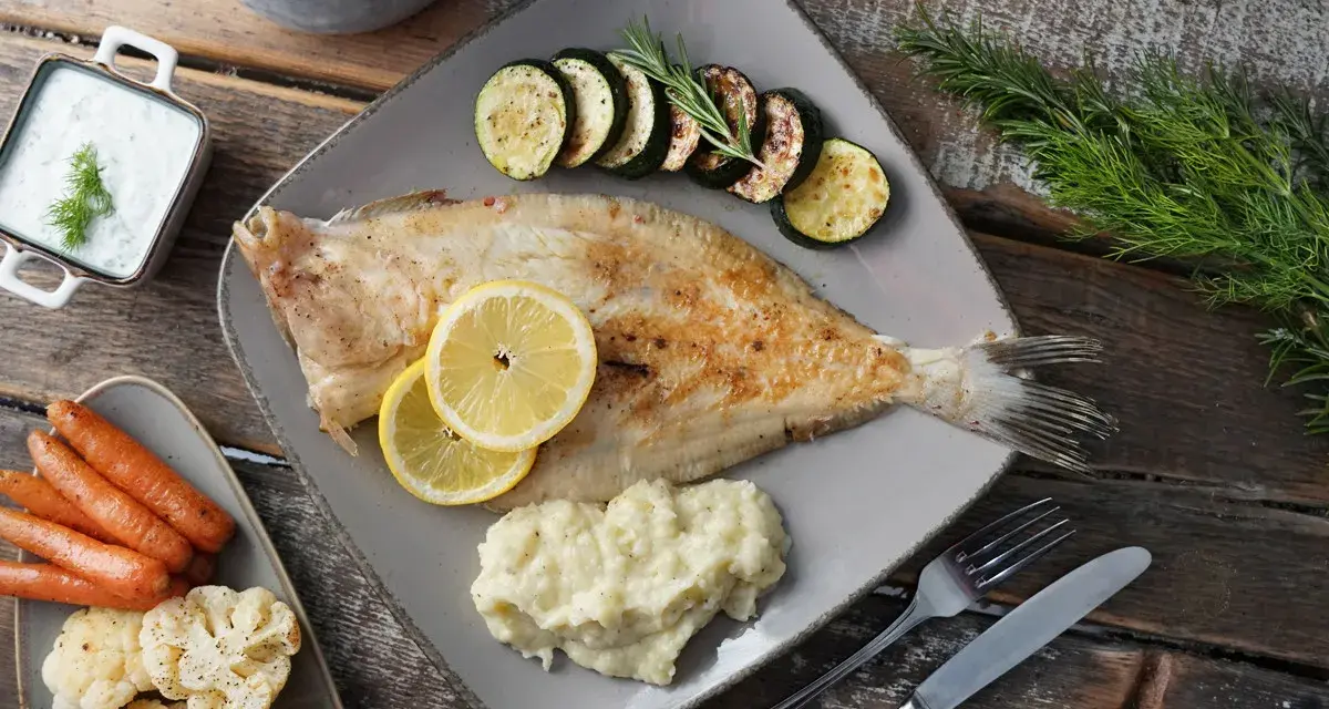 Gebratene Scholle, ein köstlicher Plattfisch, serviert mit Zucchini, Kartoffelpüree, Karotten und Blumenkohl. Zitronenscheiben und Kräuter garnieren das Gericht.