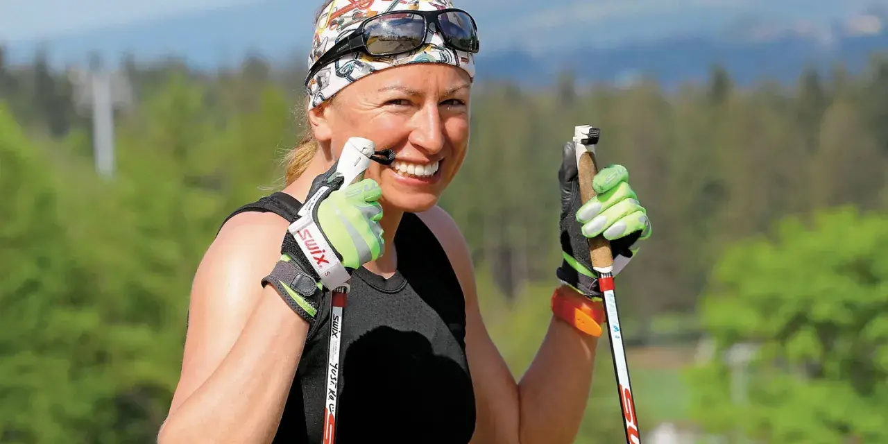 Uśmiechnięta Justyna Kowalczyk-Tekieli z kijkami do nartorolkarstwa, w sportowym stroju i opasce na głowie, gotowa do treningu.