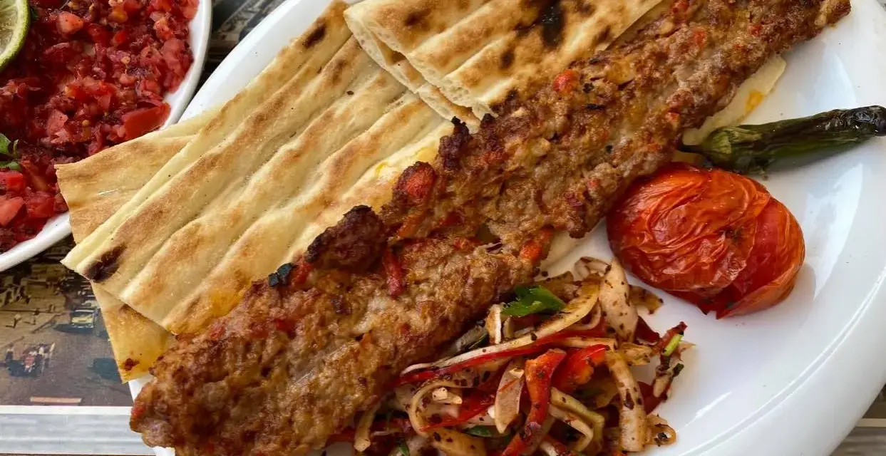 Pyszne tureckie jedzenie: kebab z mięsa mielonego, grillowany pomidor, papryka i świeże pieczywo. Obok sałatka z pomidorów.