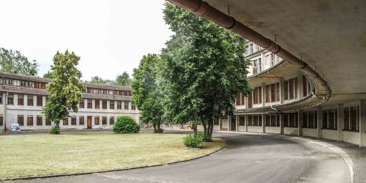 Alte Gebäude im olympisches Dorf Berlin, umgeben von Bäumen und Grünflächen. Eine Straße führt an einem geschwungenen Gebäude mit vielen Fenstern vorbei.