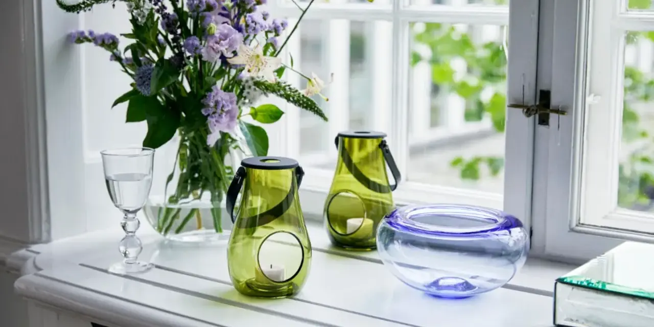 Ein Strauß lila Blumen und grüne Windlichter schmücken die Fensterbank. Ein Glas Wasser und eine blaue Schale runden die Dekoration ab.
