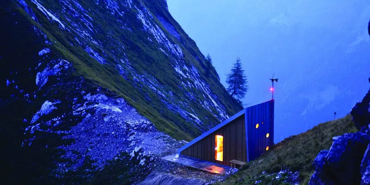Un moderno bivacco interno, illuminato da una luce calda, si staglia su un pendio montuoso al crepuscolo.