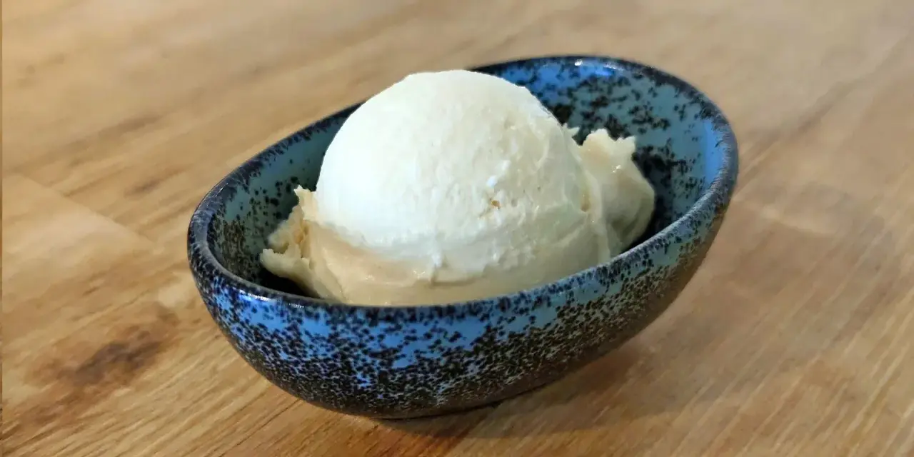 Ein Löffel cremiges Mandel Eis in einer blauen Schale auf Holztisch.