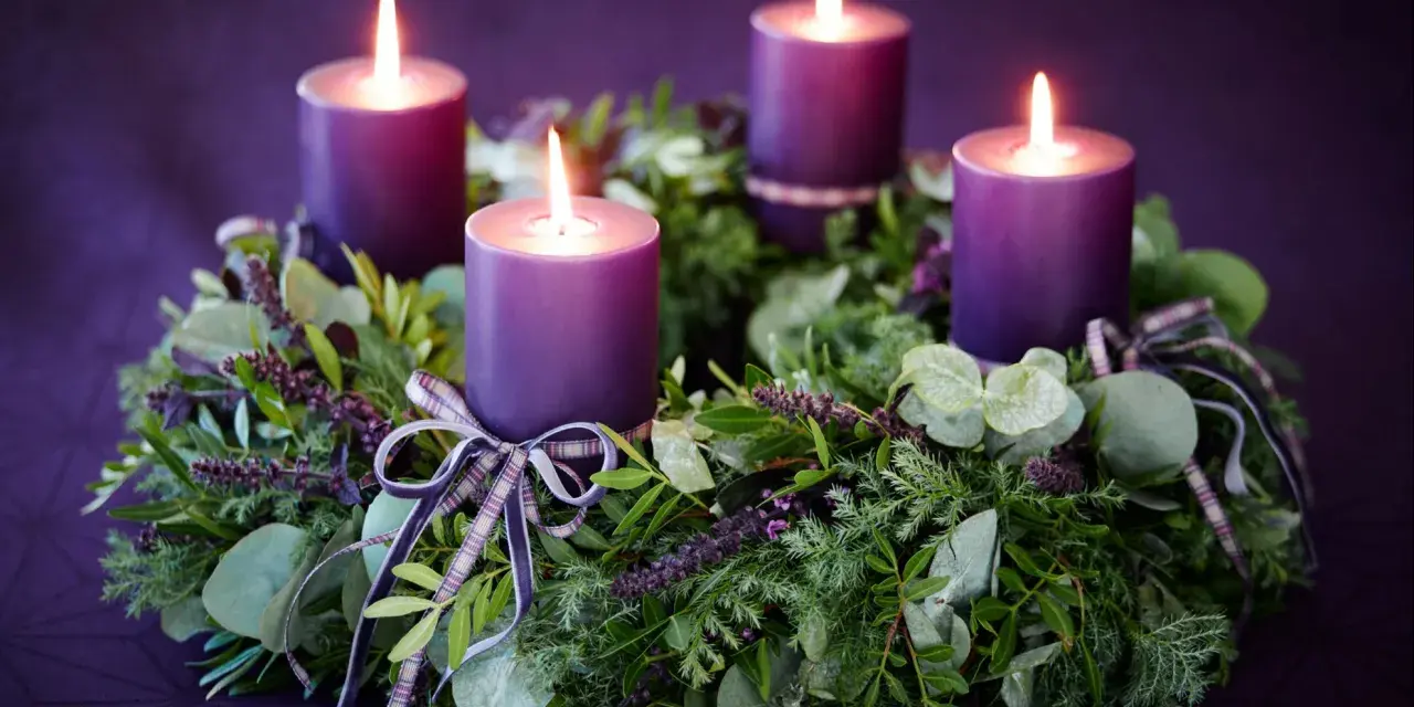 Four purple candles glow on a lush green Advent wreath, adorned with ribbons. The final candle's flame flickers, marking the 4th Advent.