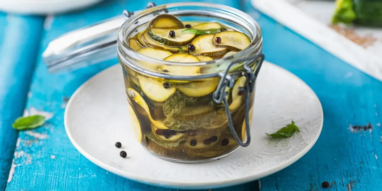 Barattolo di zucchine in agrodolce, con pepe nero e foglie di menta, su un piatto bianco appoggiato su un tavolo blu.