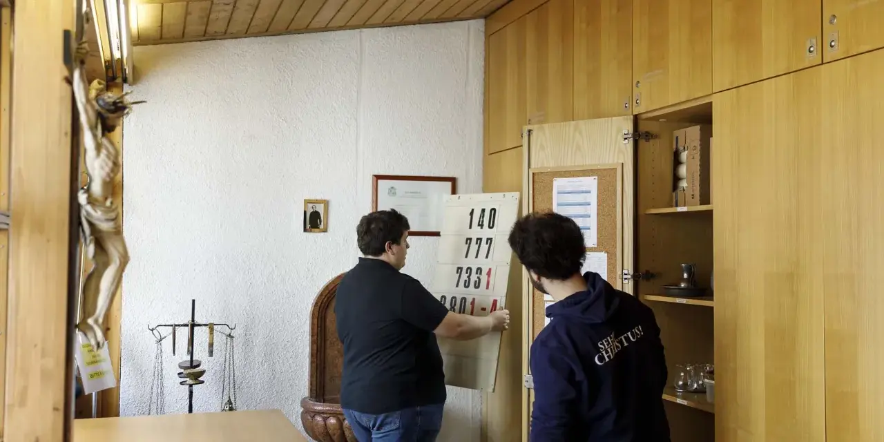 Zwei Männer im katholischen Priesterseminar hängen eine Liste mit Zahlen auf. Ein Kruzifix hängt an der Wand.
