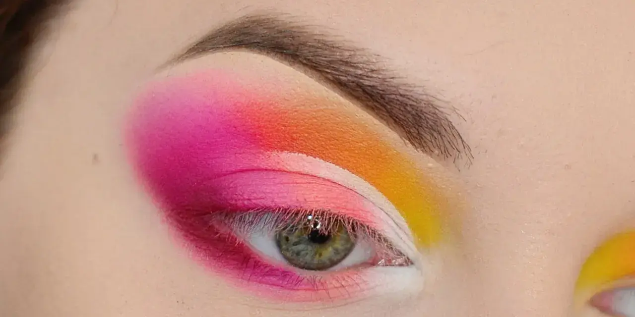 Oko z intensywnym makijażem w stylu cut crease, łączącym odcienie różu, pomarańczy i żółci.