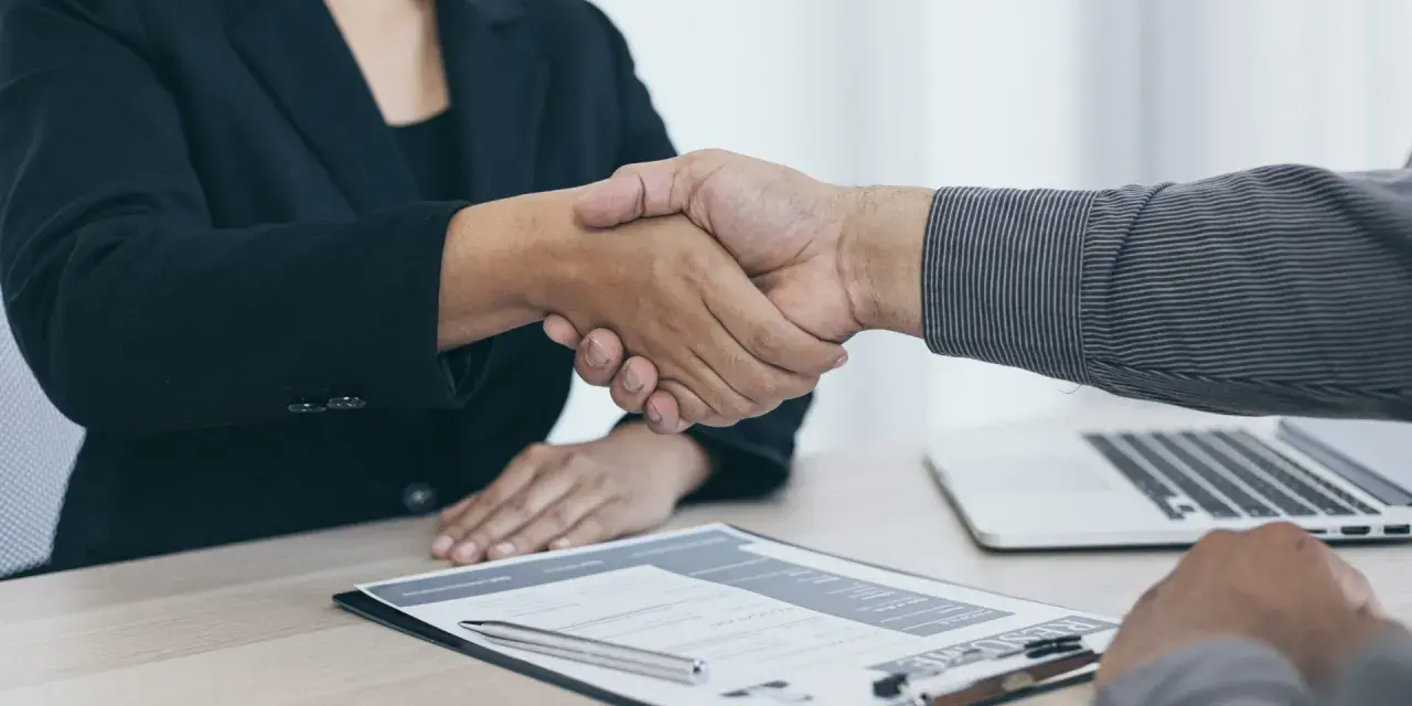 Dos personas se dan la mano sobre un escritorio con un currículum y un portátil. Es el final de una entrevista en francés.