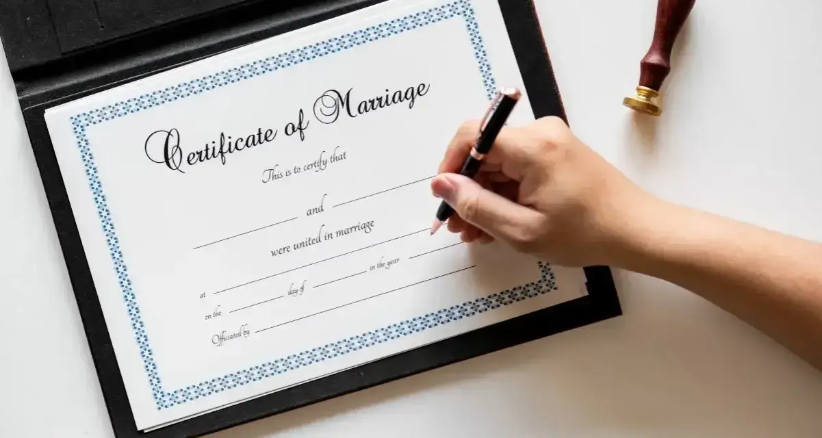 Dłoń pisze piórem na dokumencie "Certificate of Marriage", który poświadcza zawarcie związku małżeńskiego.