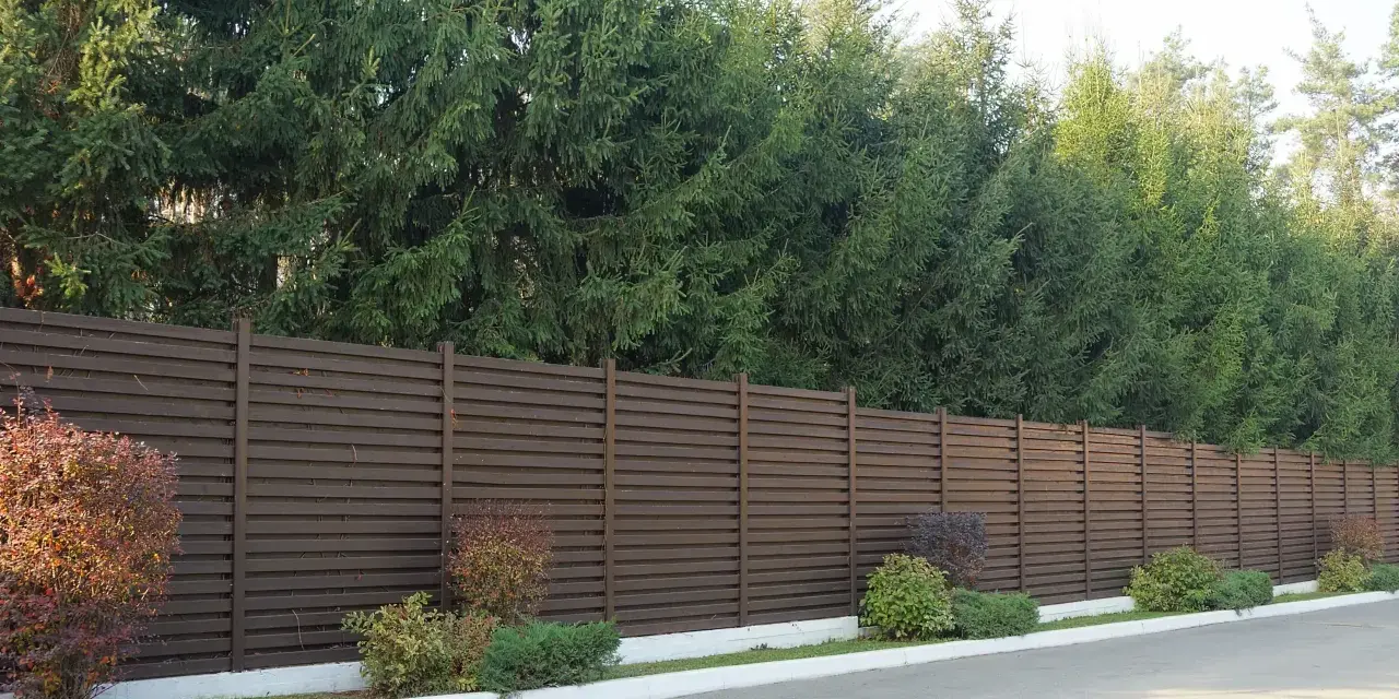 Nowoczesny, brązowy płot lamelowy to świetny sposób, by czym zasłonić płot od sąsiada. Za nim rosną wysokie świerki.