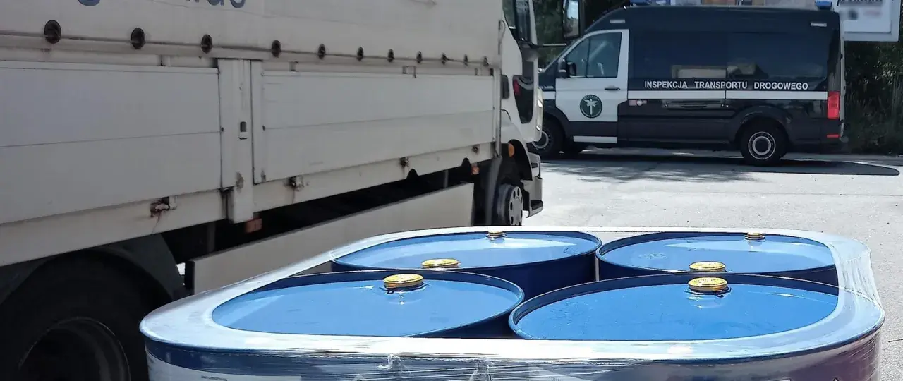 Niebieskie beczki z żółtymi zamknięciami, przygotowane do transportu towarów niebezpiecznych, obok ciężarówki i radiowozu Inspekcji Transportu Drogowego.
