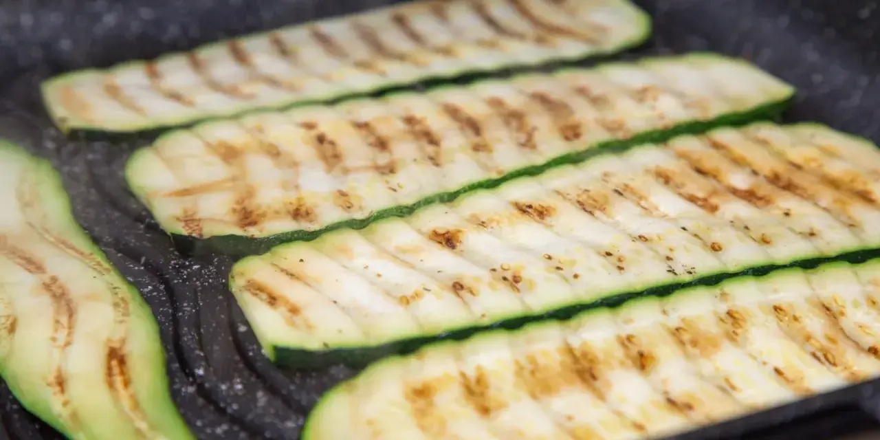 Fette di zucchine grigliate che cuociono su una piastra, con segni di grigliatura dorati e invitanti.