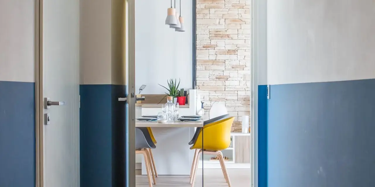 Peinture bicolore sur les murs d'un couloir et de portes, ouvrant sur une salle à manger moderne avec des chaises colorées.