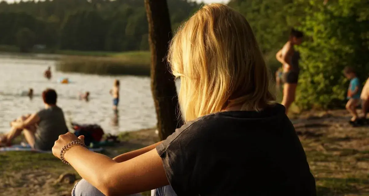 Dziewczyna z blond włosami siedzi nad jeziorem Trzechowskim, obserwując ludzi kąpiących się w wodzie.