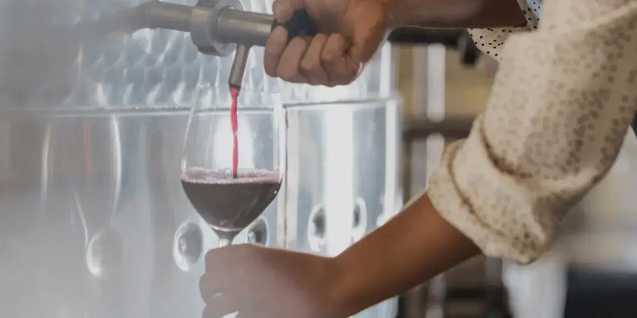 Rött vin hälls upp i ett glas från en stor tank. En hand vrider på kranen, kanske för att kontrollera tillsatser i vin.