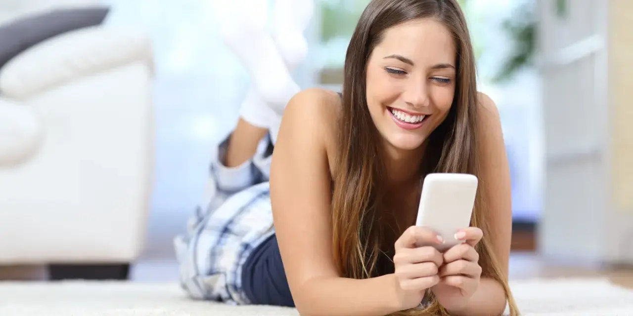 Uśmiechnięta dziewczyna w piżamie leży na dywanie, przeglądając telefon. Może szuka nowych znajomości na Tinder randki.