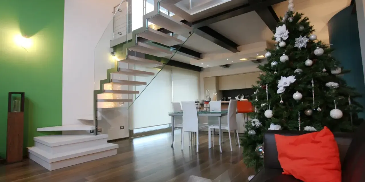 Un moderno soppalco casa con scala in vetro e legno, albero di Natale addobbato e un divano con cuscino arancione.