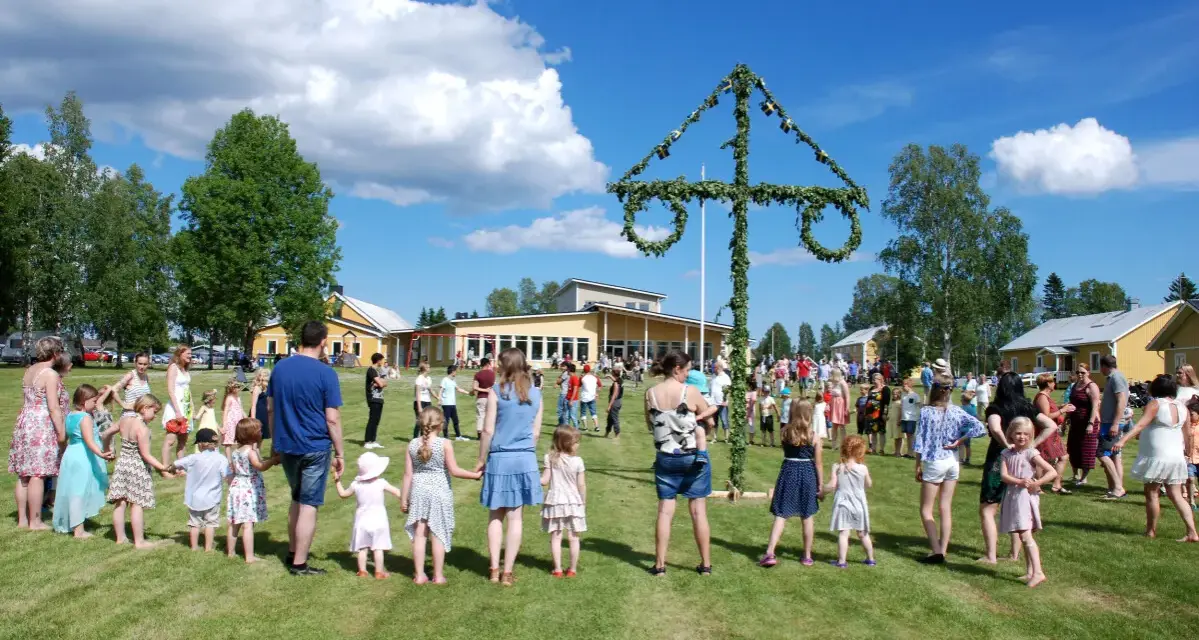 Många människor, både vuxna och barn, håller varandra i händerna och dansar runt en midsommarstång. De firar midsommar i en grönskande park med gula byggnader i bakgrunden.