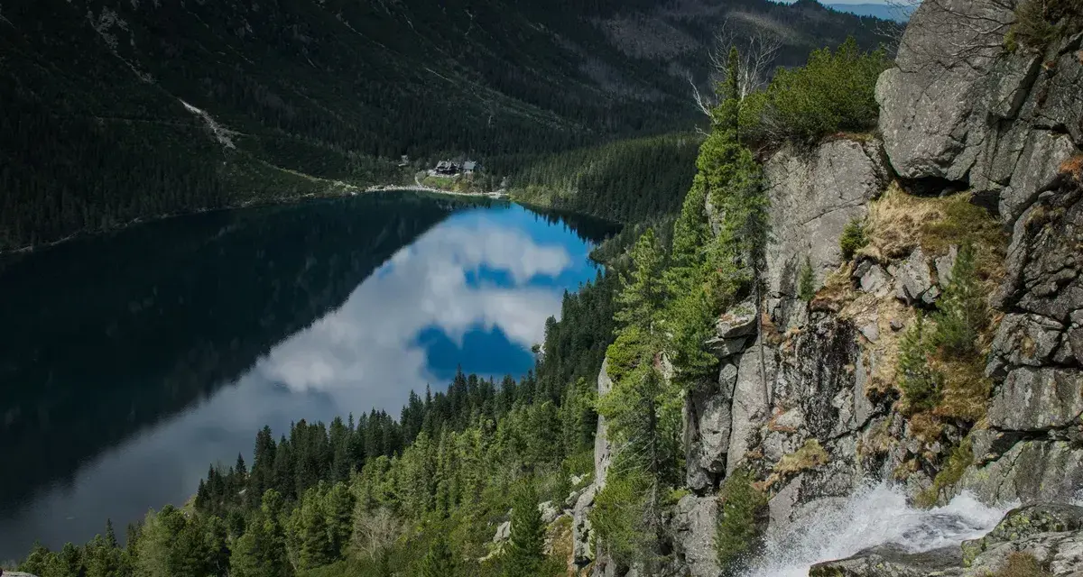 Widok z góry na Morskie Oko, odbijające chmury. Trasa turystyczna prowadzi przez skaliste zbocza z wodospadem.