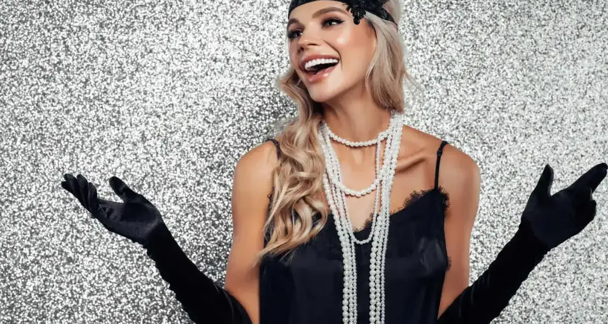Uśmiechnięta kobieta w czarnej sukience i długich rękawiczkach, z perłami i opaską na głowie, uosabia szykowną elegancję lat 20. stylizacje.