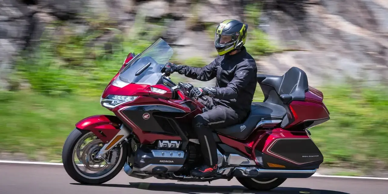 Mężczyzna w kasku jedzie lśniącym, czerwonym motocyklem uniwersalnym. W tle rozmyte zielone wzgórza i skały.