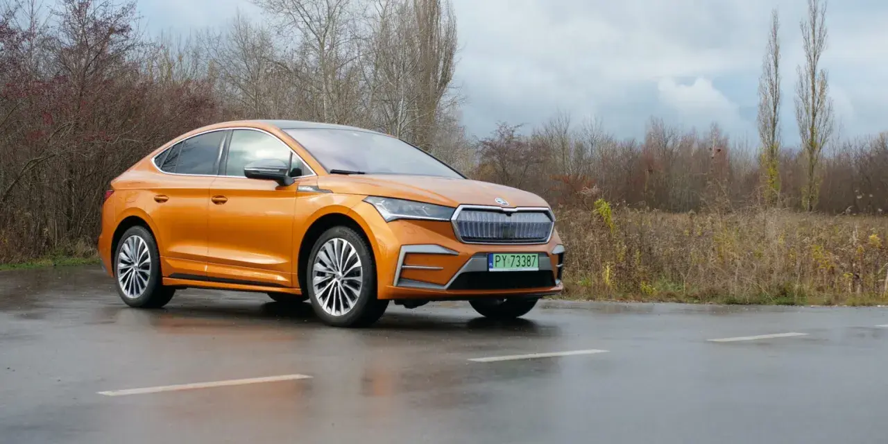 Pomarańczowa Skoda Enyaq Coupe na mokrej drodze, zarośla w tle.